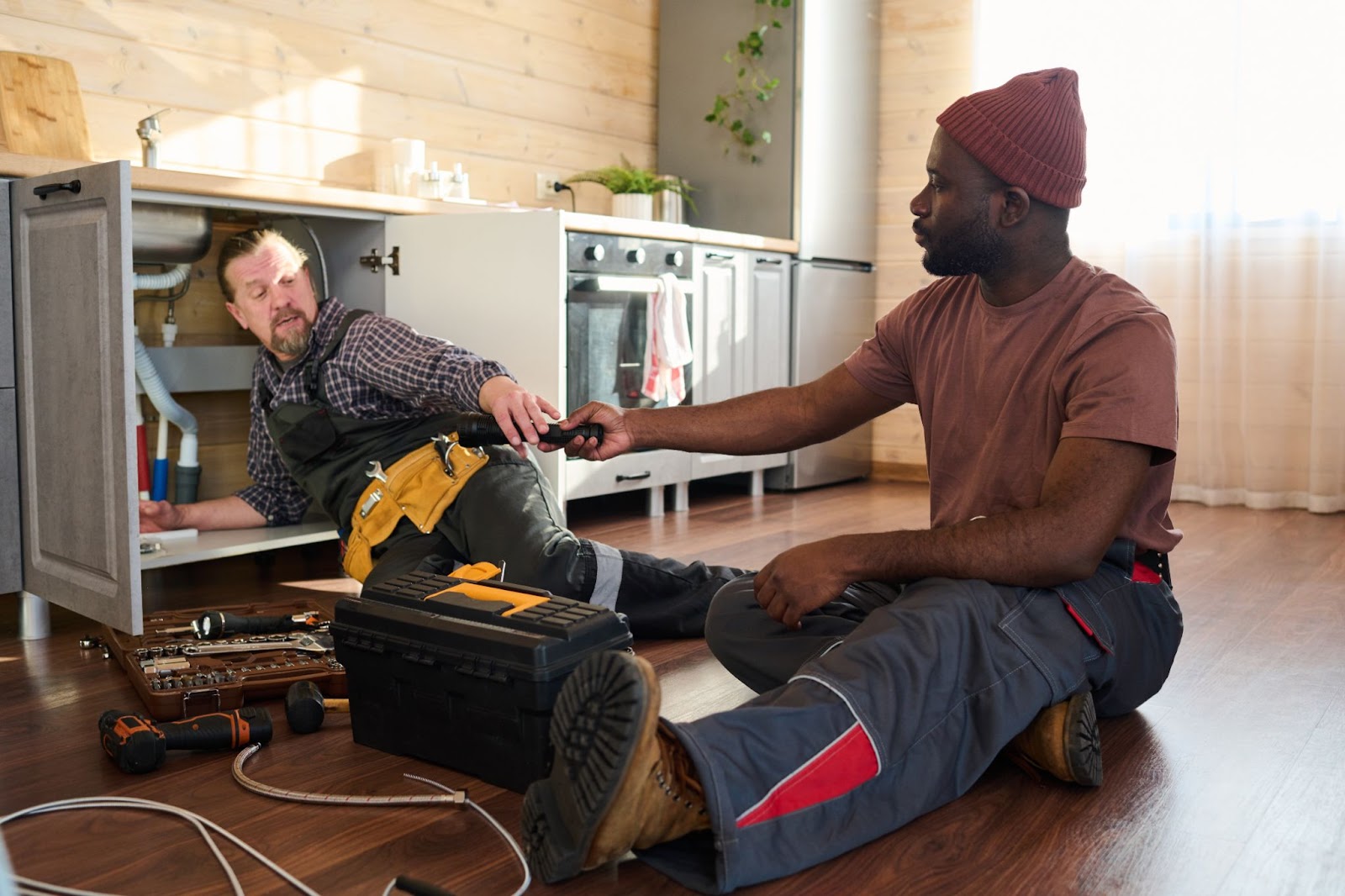 A handyman in workwear hands a tool to his experienced colleague while making plumbing repairs.