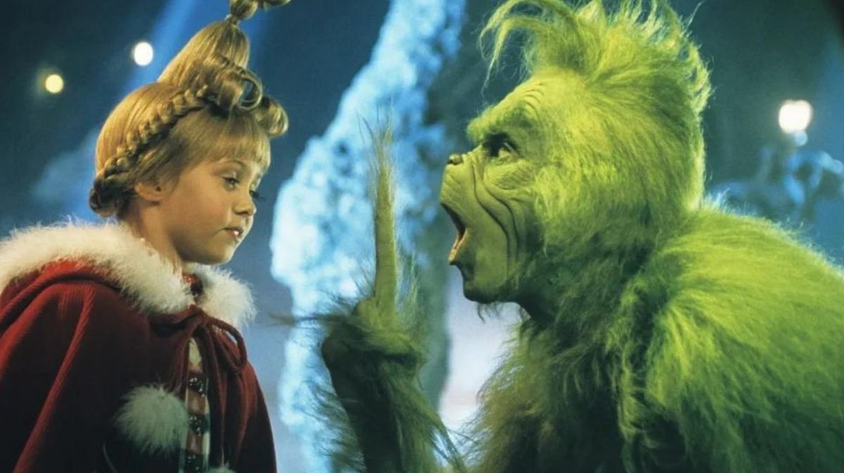 El Grinch verde y una niña rubia vestida de rojo conversan en una escena navideña.