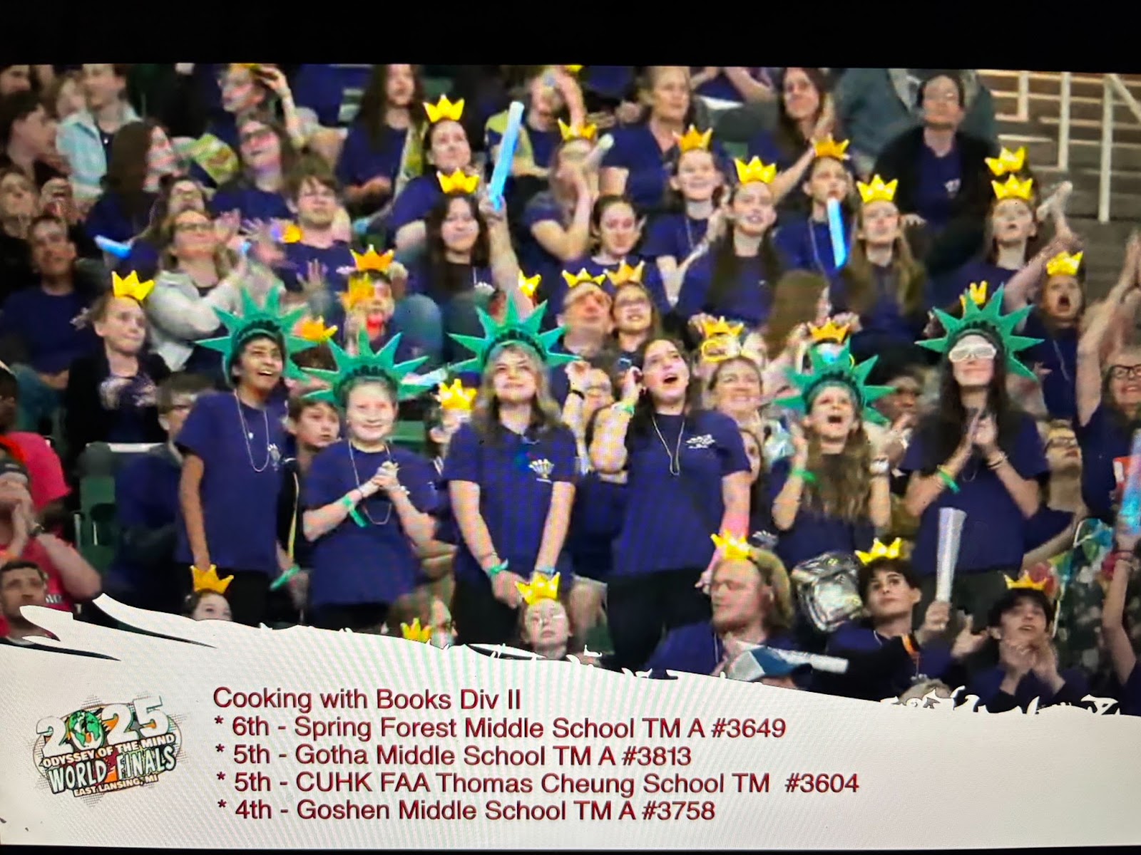 Students stand and clap in a crowd as they are shown on a video board. They are wearing statue of liberty style hats and matching Odyssey of the Mind shirts. 