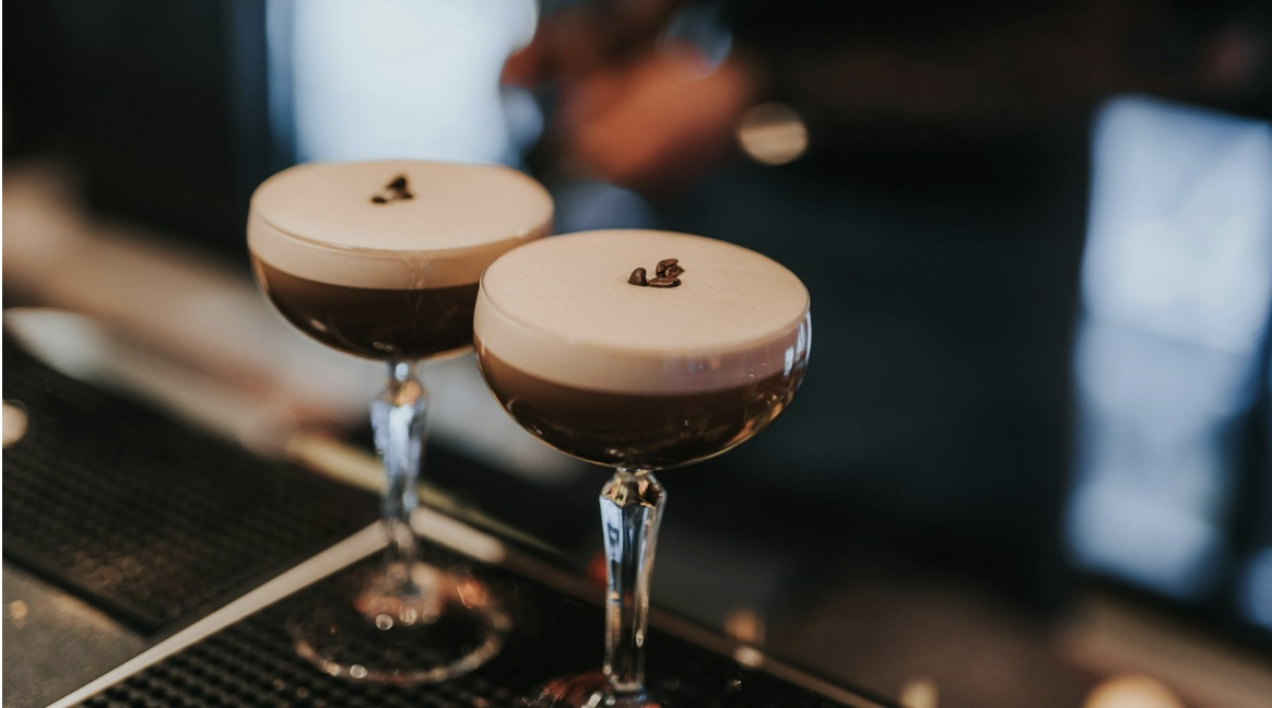 Two Espresso Martinis with three coffee bean garnishes are on a black bar mat