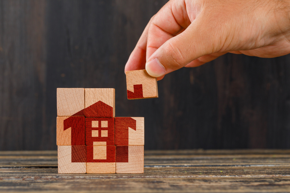 A hand carefully places a small wooden block into a puzzle, creating the image of a red house with windows and a door, as if an architect crafting Vastu design, all set against a wooden background.