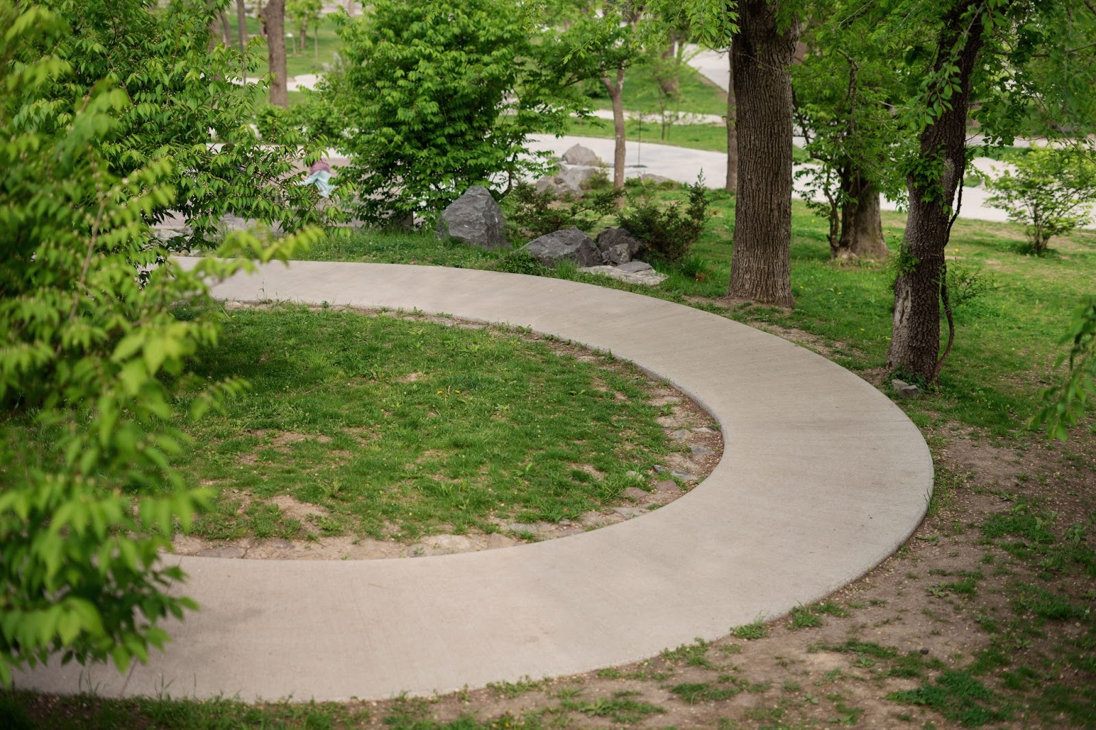 A winding concrete sidewalk surrounded by trees, grass, and rocks.