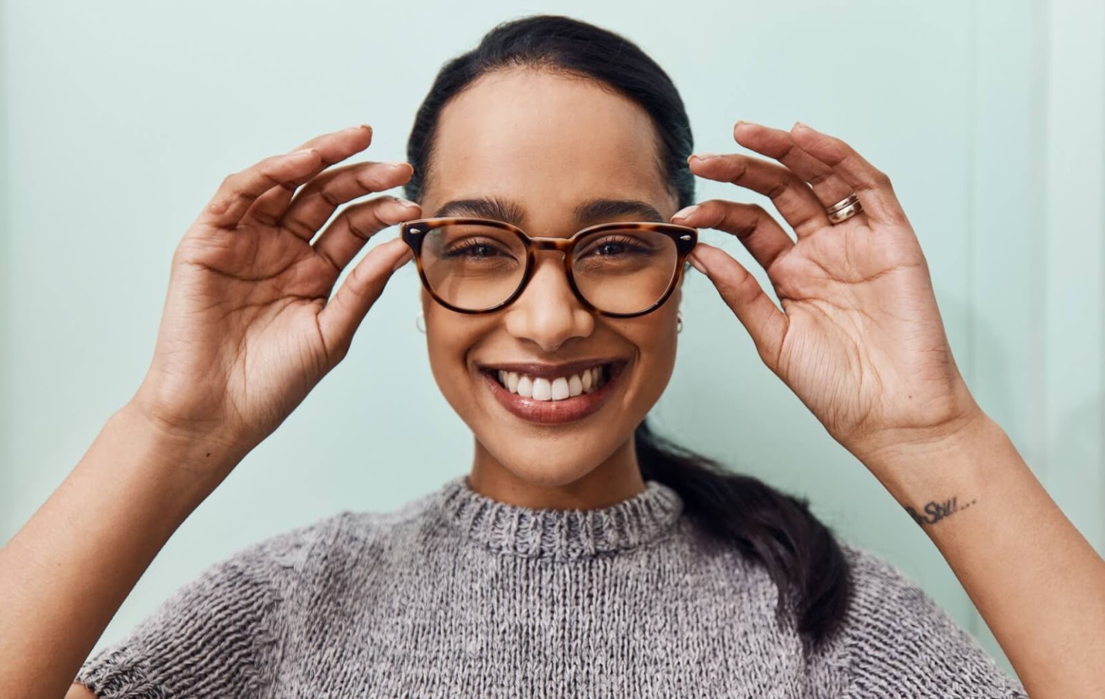 A young person wearing a grey knit shirt smiles widely as they put on a pair of glasses to correct their 20/30 vision.
