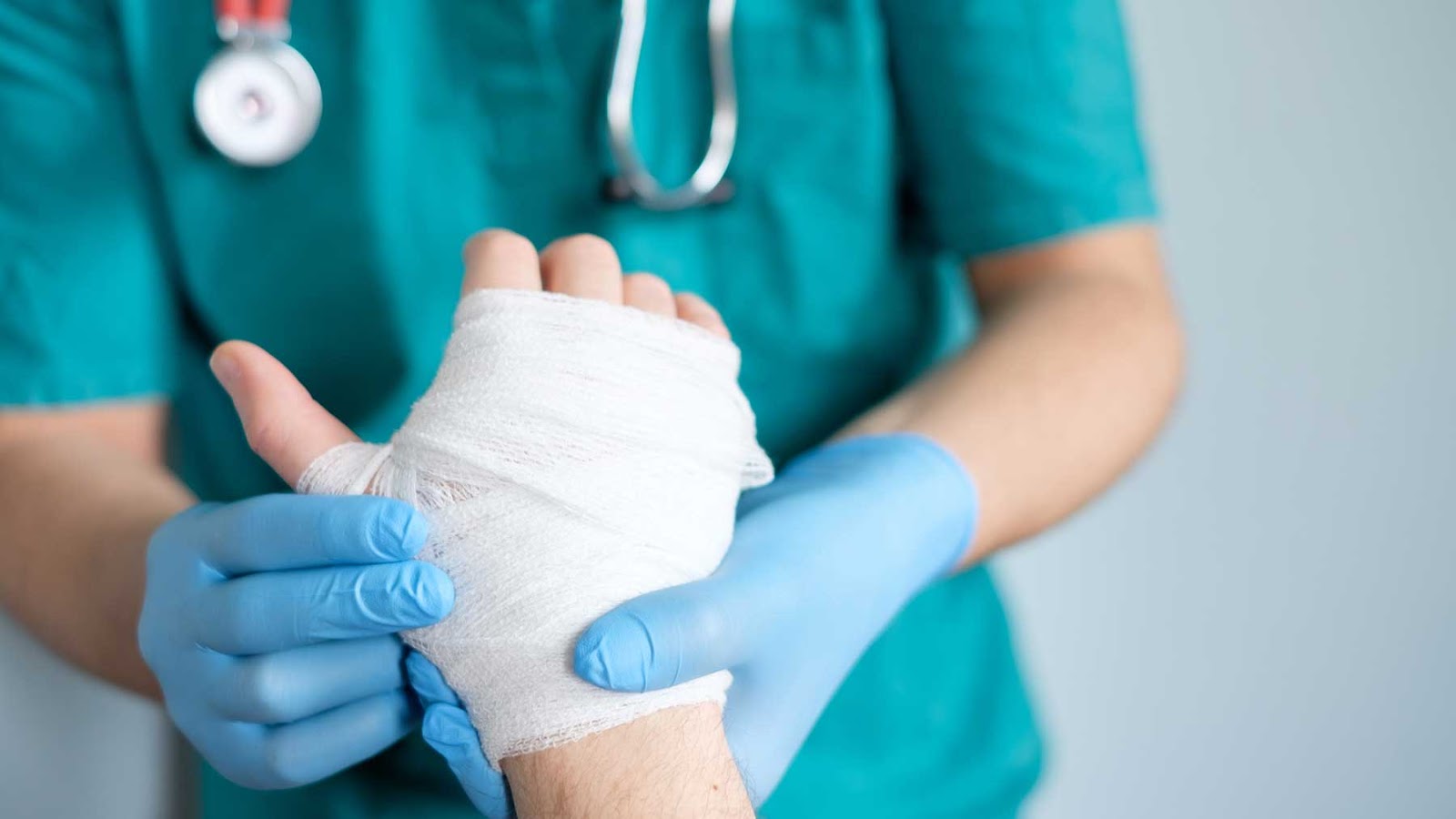 A healthcare professional wearing blue latex gloves is carefully wrapping a patient's injured hand with a white bandage. 