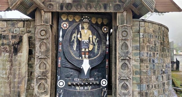 Ornate wooden door with traditional carvings and a central figure holding a spear and shield, emblematic of Nagaland architecture. The door showcases motifs and symbols, flanked by stone walls with tile patterns.