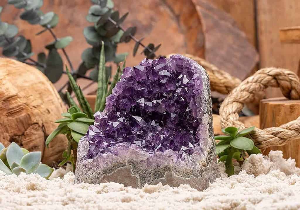 A geode with a cluster of amethyst crystals inside. The geode is placed on a surface covered with sand, surrounded by decorative elements such as green succulent plants, a rope, and wooden objects. The amethyst crystals have a deep purple color, typical of this variety of quartz.