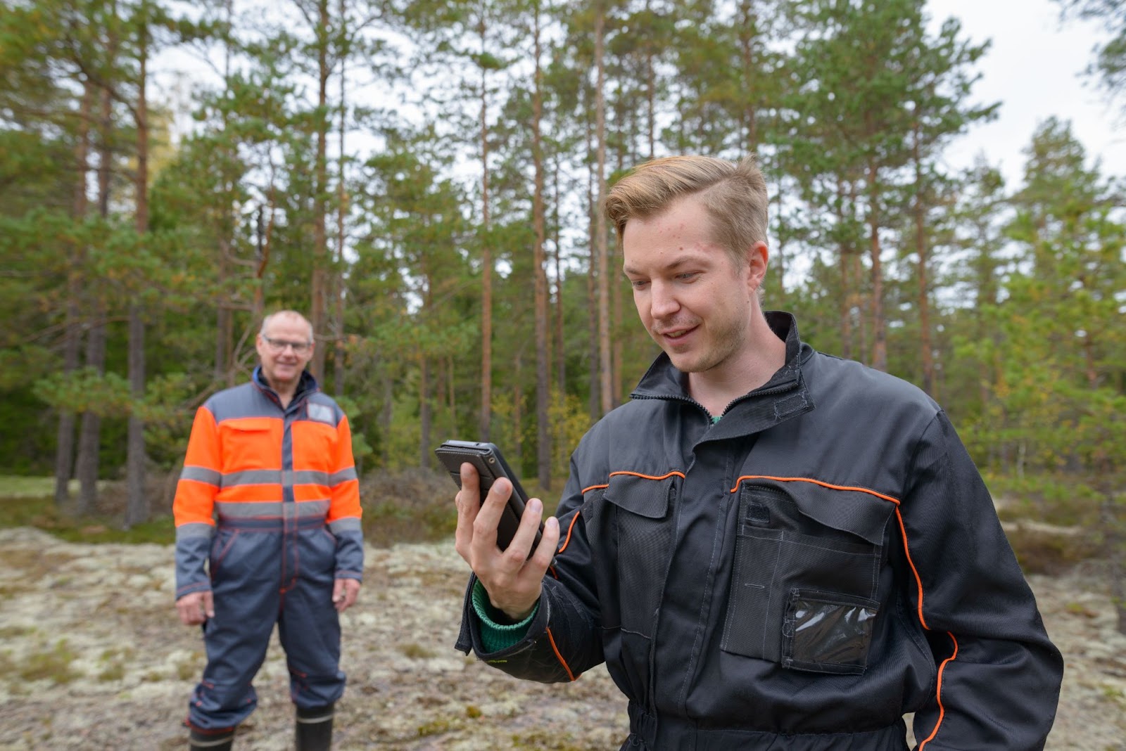 A young forestry technician in black holds a mobile phone in the forest, with a mature forester in safety gear smiling in the background. Both look pleased with the use of advanced tools.