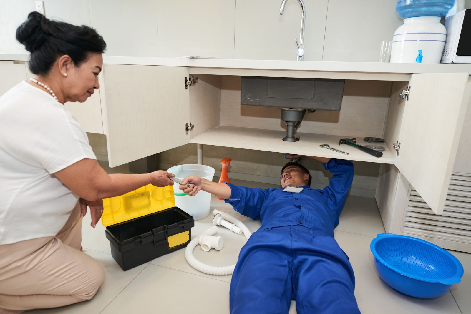 A woman kneeling next to a plumber as he works.