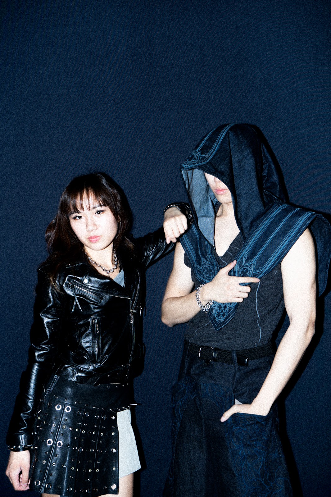 Two models stand against a midnight blue background. The female model on the left stands in a punk-inspired leather jacket and studded skirt, next to a male model wearing a hooded avant-garde look. 