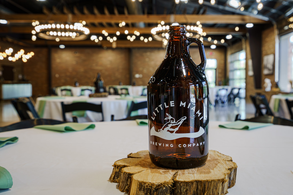 Close-up of a brown glass growler from Little Miami Brewing Company placed on a rustic wooden coaster in a well-lit, elegant event space with round tables and green napkins in the background.