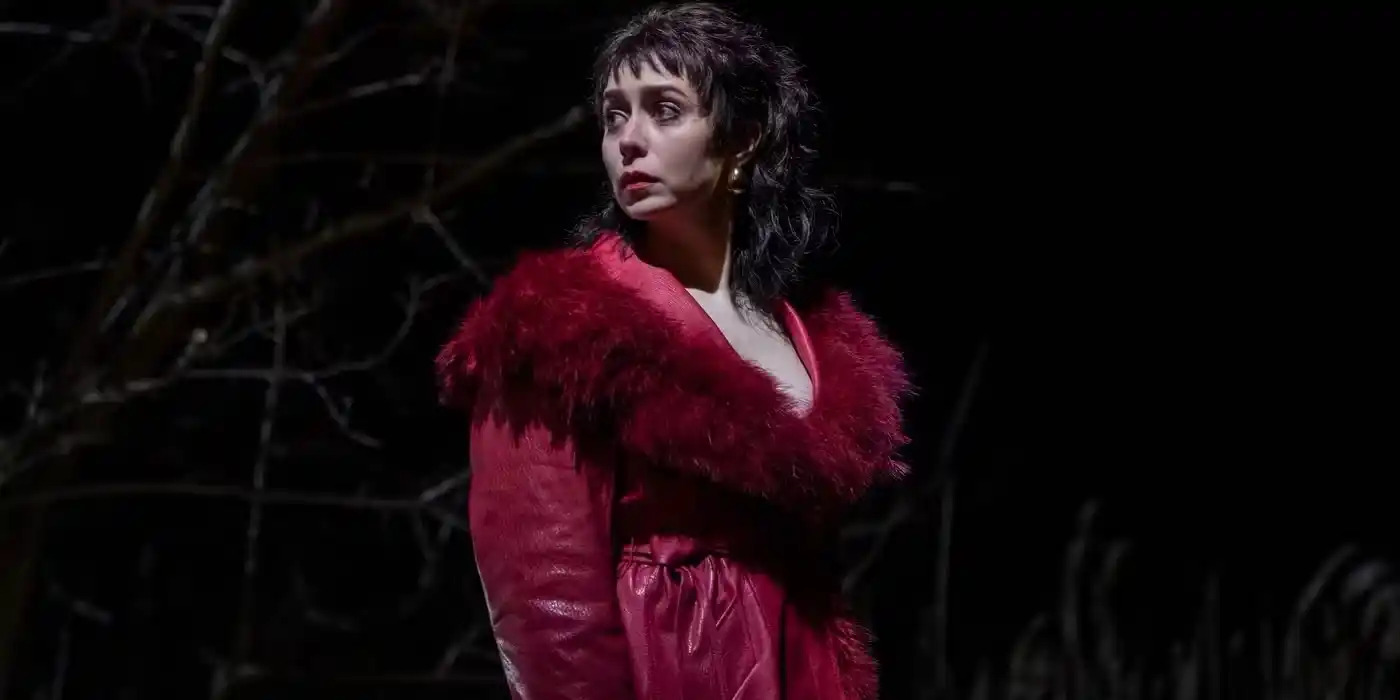 Mujer de cabello corto y oscuro, con un vestido rojo y una chaqueta con detalles de plumas, mirando hacia un lado en un entorno oscuro con árboles al fondo.