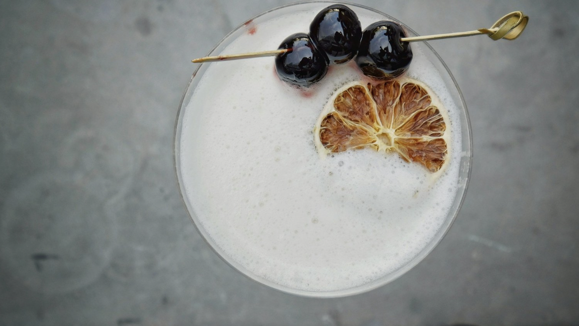 The top of a whiskey sour cocktail has three cherries on a bamboo toothpick and a dehydrated lemon wheel