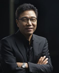 This contains an image of lee soo man on a black background putting on a black suit and folding his hands