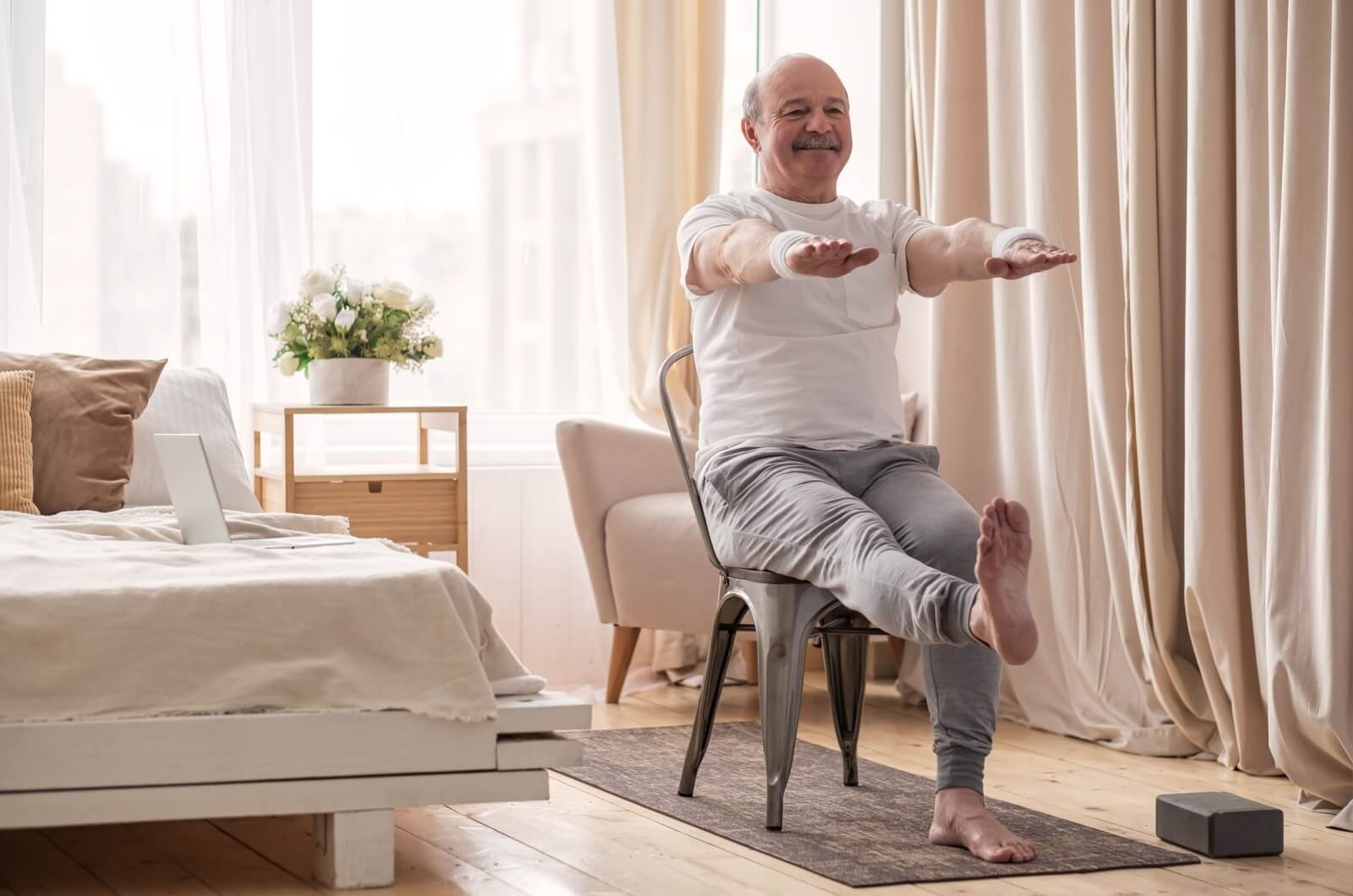 A smiling older adult performing leg lifts while sitting on a chair in a bedroom.