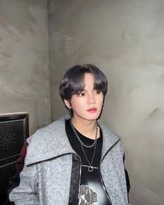 This contain an image  UP10TION's Hwanghee with a  black hair wearing a gray jacket and necklace, standing in front of a wall