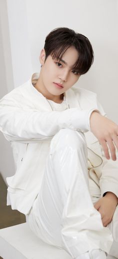 This contains an image of Doyoung sitting on top of a white bench wearing all white clothing and looking at the camera