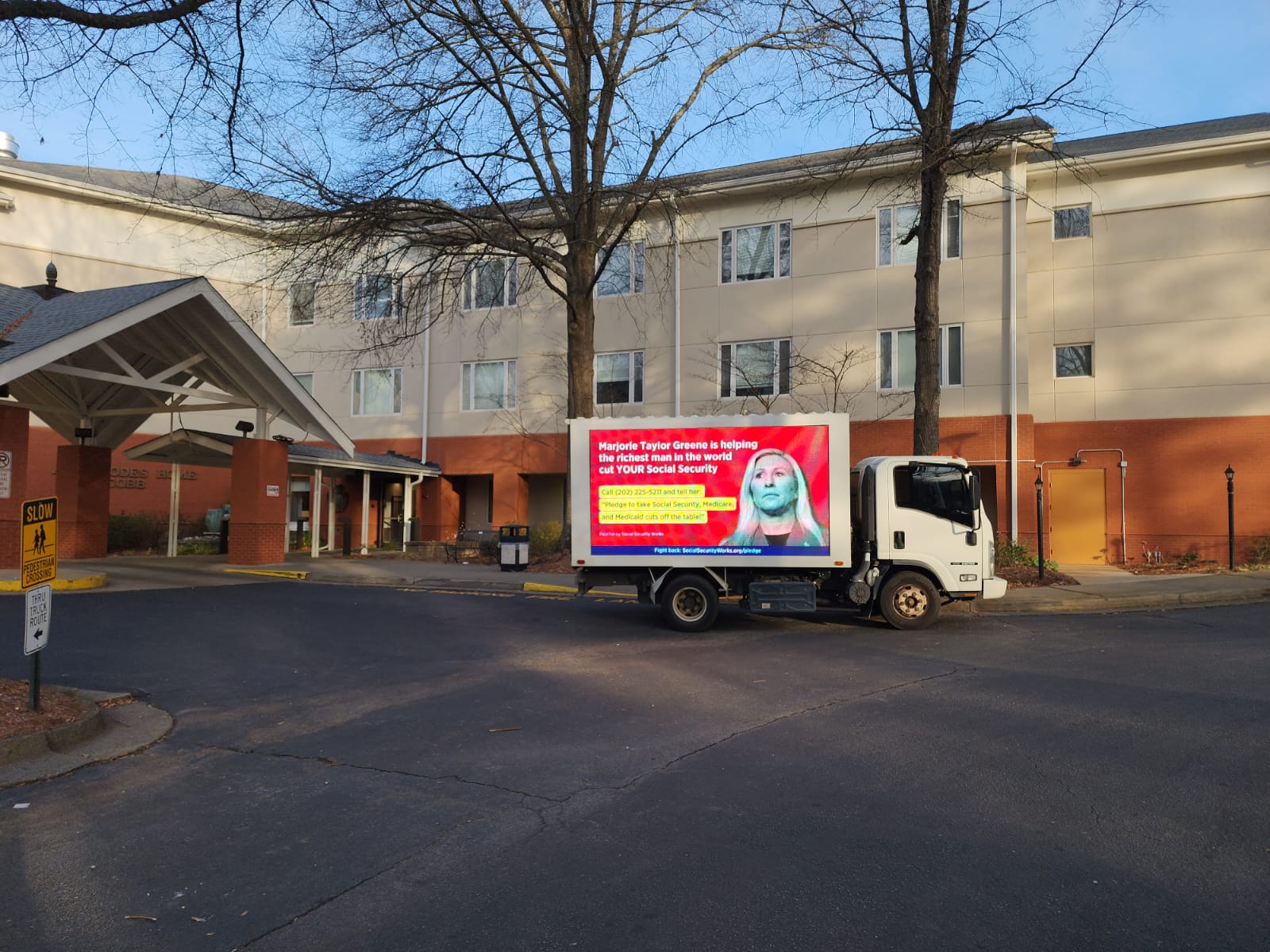 A billboard truck is parked outside of a Georgia senior center. The billboard displays Rep. Marjorie Taylor Greene's face with the text 'Marjorie Taylor Greene is helping the richest man in the world cut YOUR Social Security. Call (202) 225-5211 and tell her to pledge to take Social Security, Medicare, and Medicaid cuts off the table. 