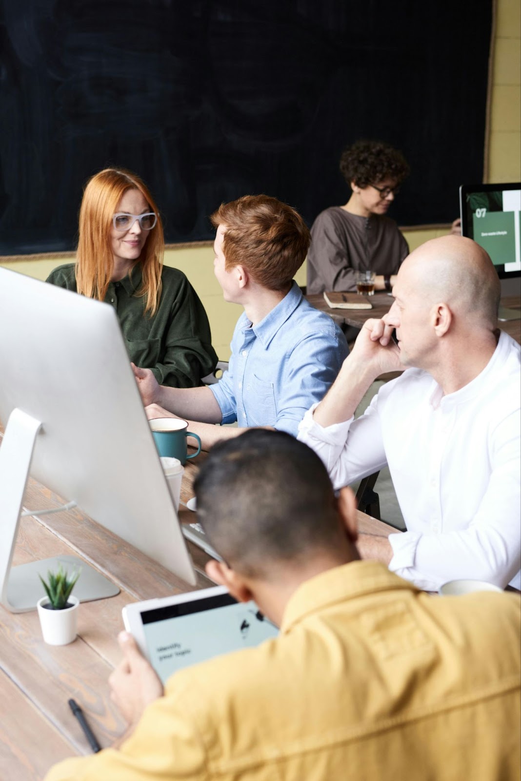 A group of people working and discussing around a table, using tablets and desktop computers.