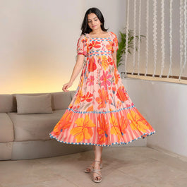 Woman wearing a flowy pink maxi dress with bold orange and red floral prints, featuring puff sleeves and tiered detailing.