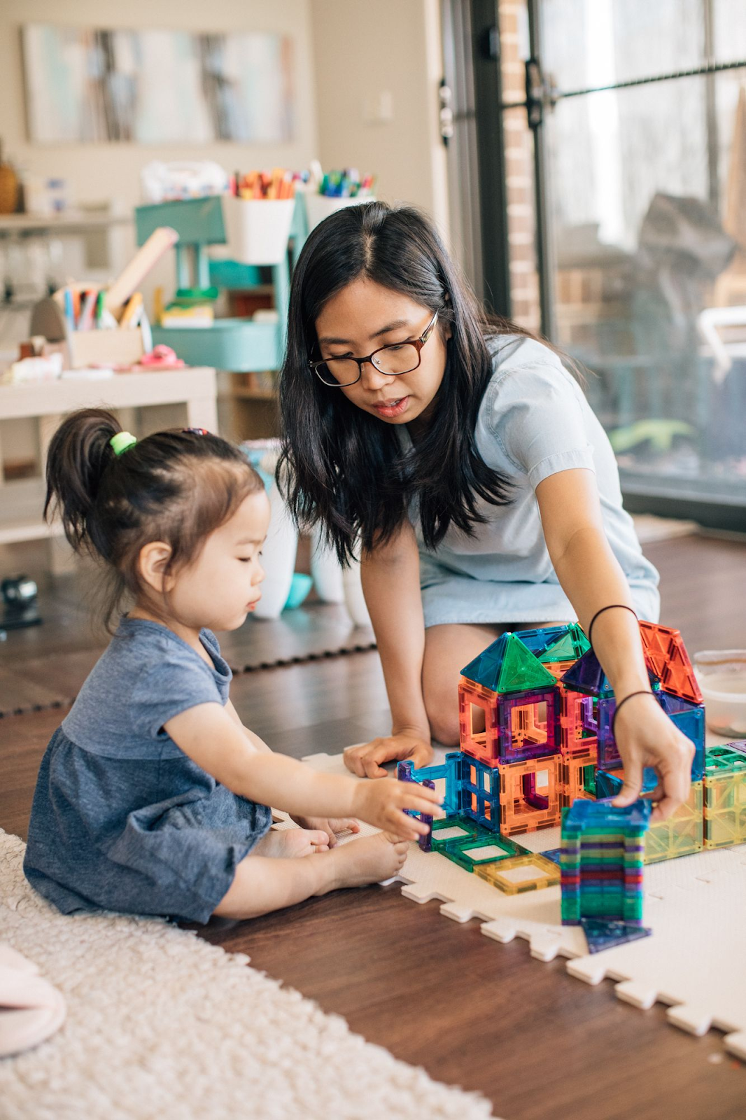 A parent and toddler girl are engaged in a playful session with magnetic tiles, with the parent showing her how to stack the tiles by placing one piece on top of another. The toddler is focused and excited, enjoying the hands-on learning experience as they create colourful designs together.