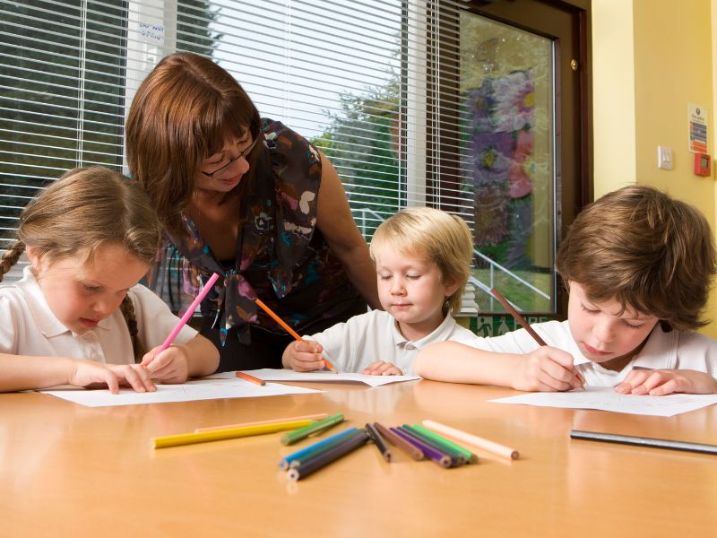 A teacher assists three young children drawing at a table.