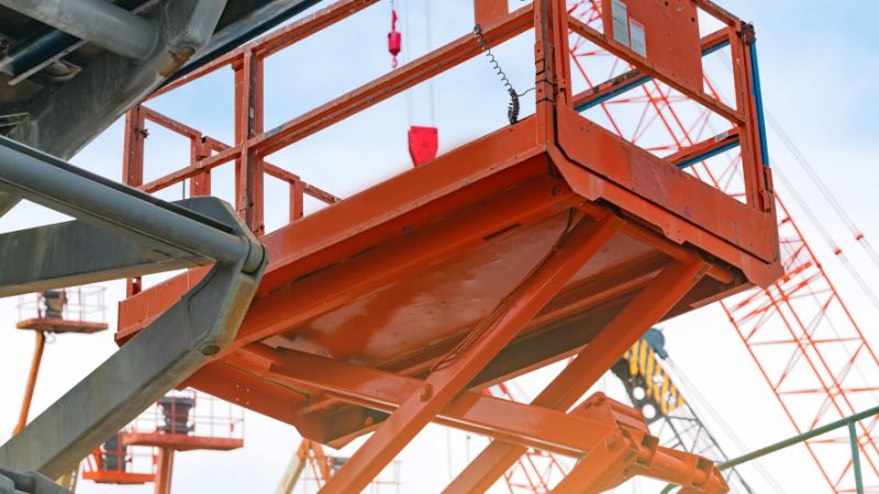 Plataforma elevatória tipo tesoura laranja com guindastes ao fundo contra um céu azul.