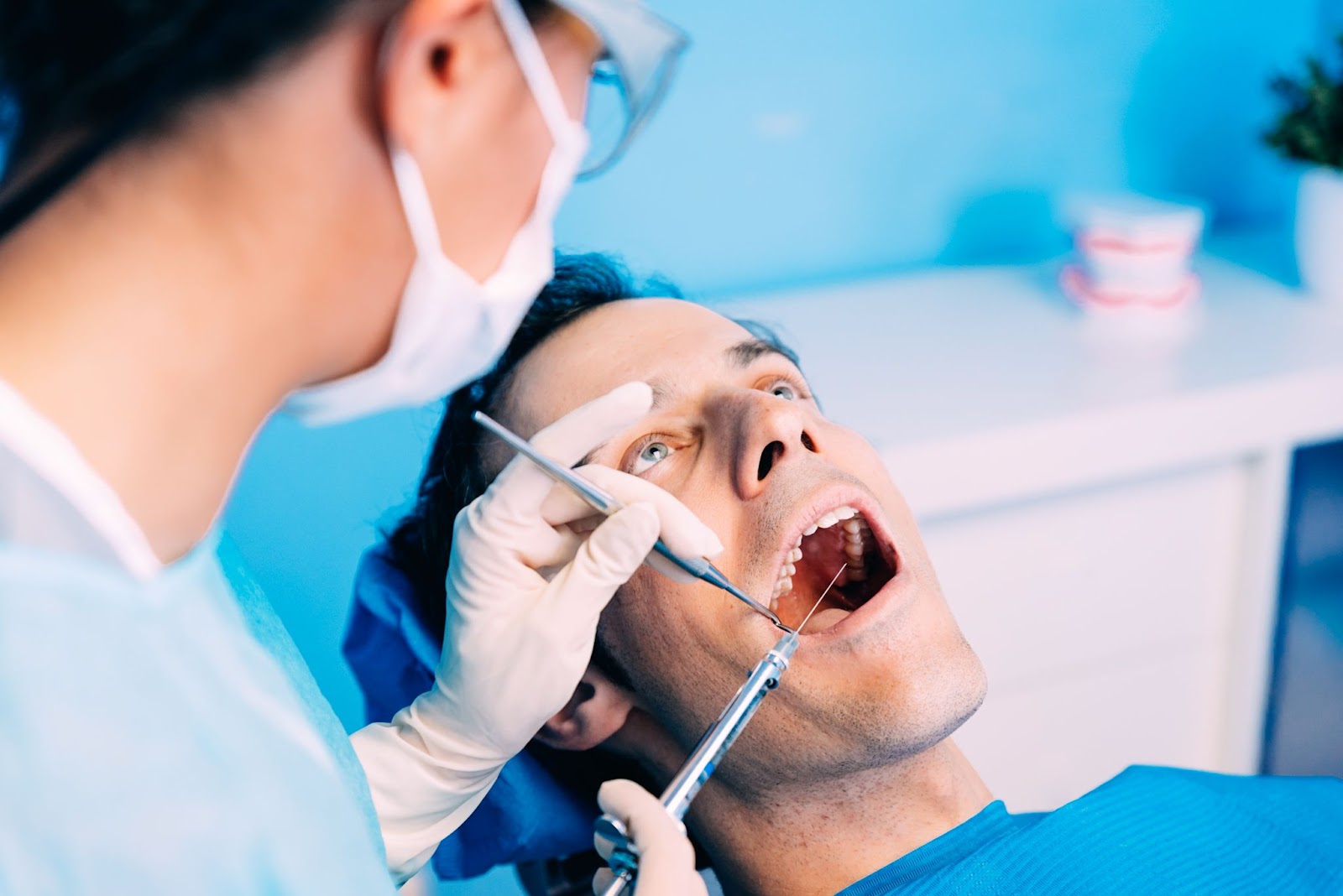 A close-up of a patient undergoing a dental procedure, with gloved hands of a nurse holding various dental tools inside the patient's mouth. The patient looks relaxed as the dentist works, with a clean and professional blue-toned background emphasizing a sterile and clinical environment.