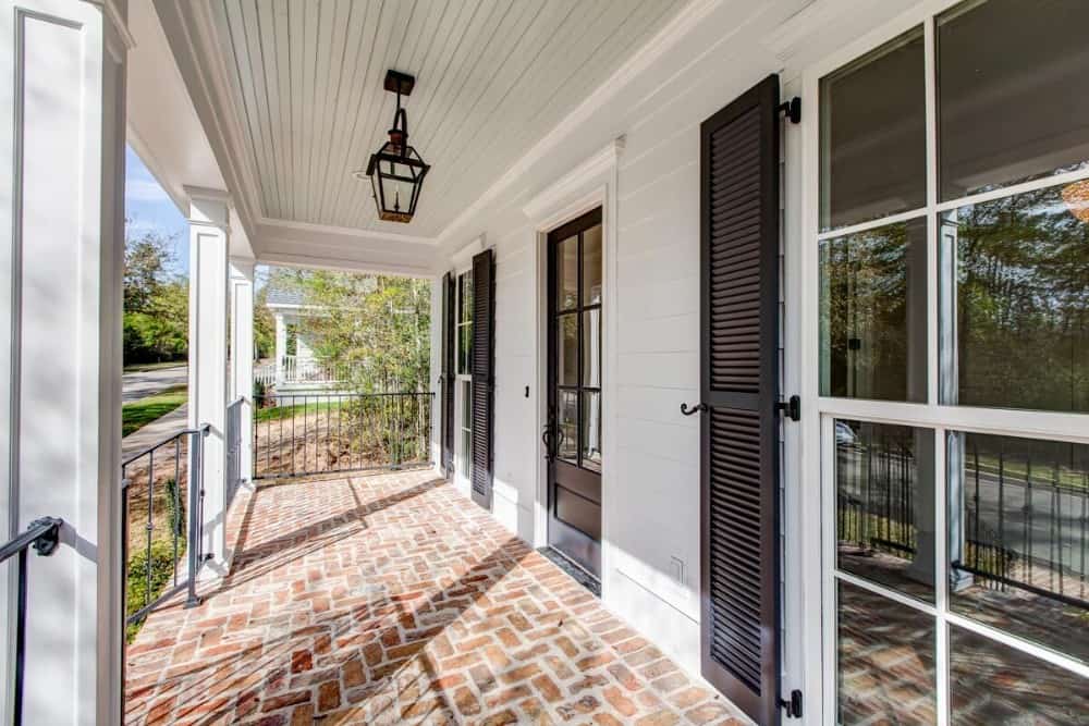 Covered front porch with white columns, brock flooring, shuttered windows, and a glazed entry door.