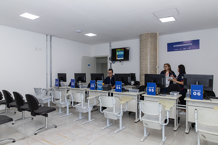 A imagem mostra um ambiente de atendimento ao público, possivelmente em uma farmácia ou setor de saúde. O espaço é composto por um balcão de atendimento com seis guichês numerados de 01 a 06, cada um equipado com monitores de computador e cadeiras para funcionários e clientes. Há funcionários uniformizados no ambiente: um deles está sentado no Guichê 04, enquanto outros dois estão em pé, conversando ou organizando algo próximo ao Guichê 06.  Na parede ao fundo, há uma placa azul com a palavra "ATENDIMENTO", indicando que se trata de uma área destinada ao suporte ou serviço ao público. Também há uma televisão na parede, exibindo algum conteúdo informativo. O ambiente é bem iluminado, com paredes brancas, piso claro e um pilar central.  À esquerda da imagem, há uma fileira de cadeiras pretas, possivelmente destinada aos clientes aguardando atendimento. O local parece ser organizado e projetado para oferecer conforto durante o serviço.