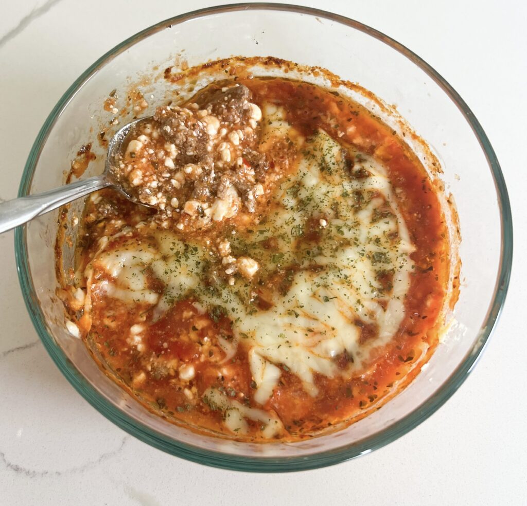 This healthy cottage cheese lasagna bowl is gluten free, high protein, low carb, keto, and single-serve. In a large glass bowl is the lasagna after baking - red marinara sauce bubbly, cottage cheese texture, herbs, melted cheese, and ground beef poking through. It looks like a classic lasagna but in the bowl. 