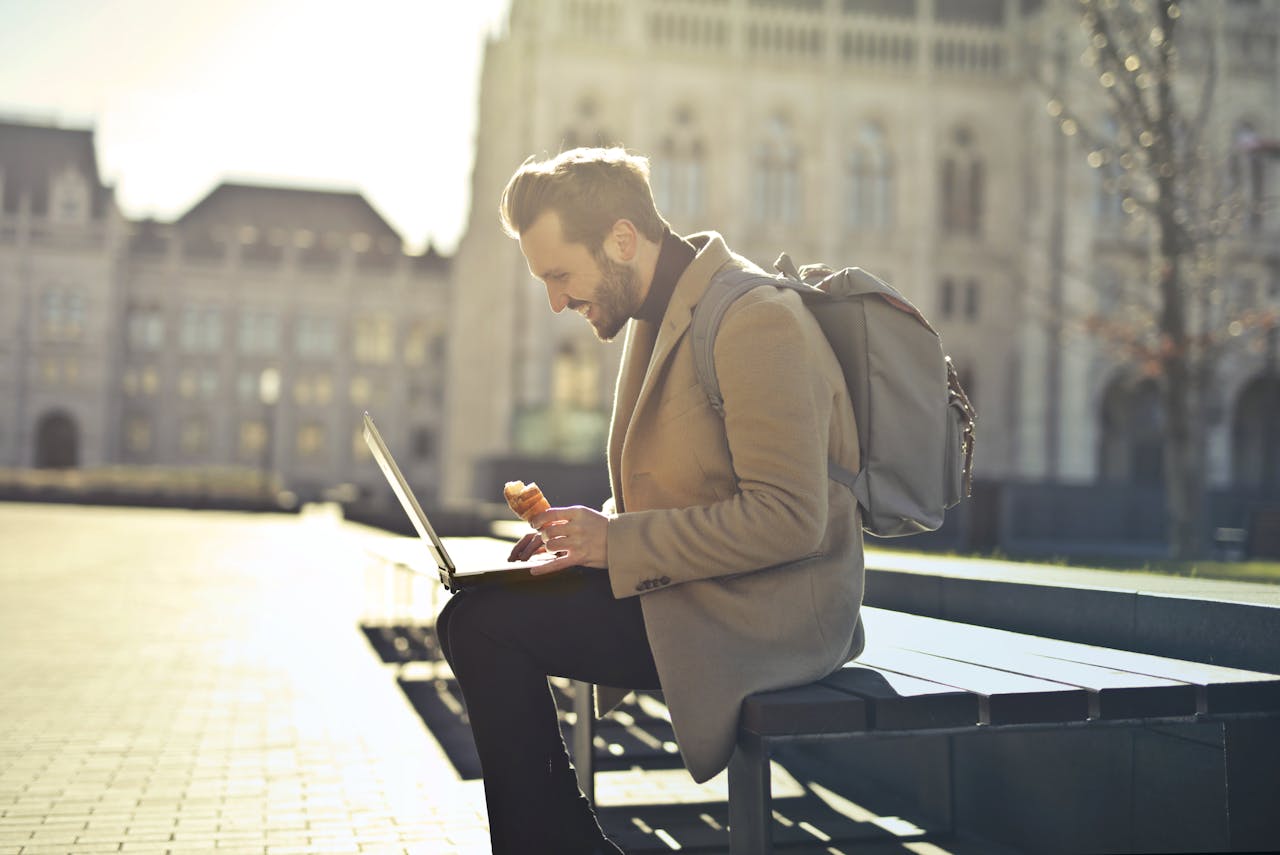 man in brown coat, sitting on a bench with a laptop on his lap