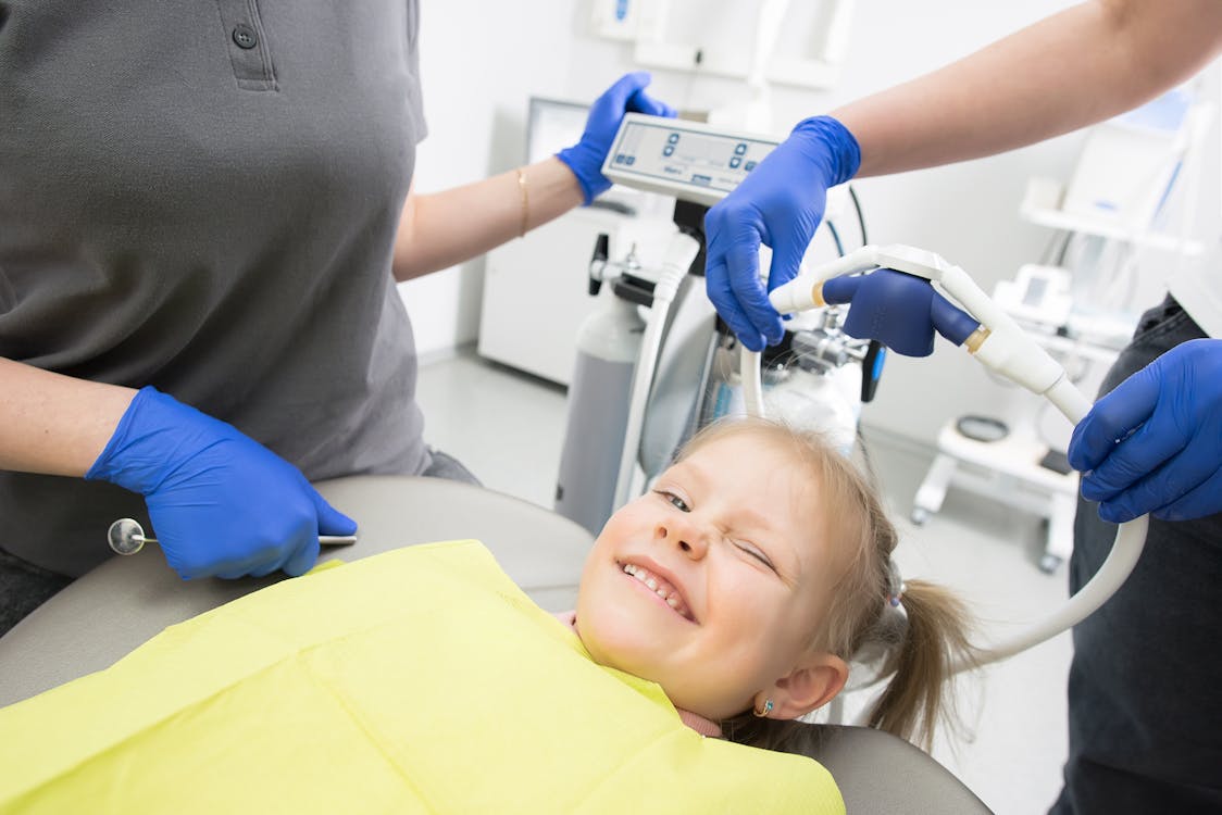 Free A cheerful child winks while sitting in a dental chair, surrounded by dental professionals. Stock Photo