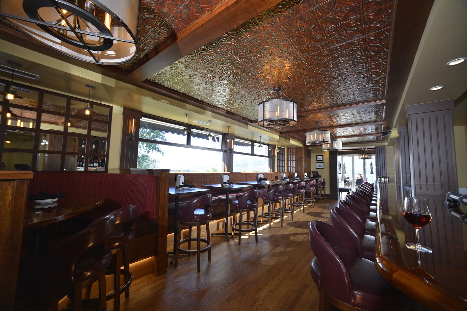 A long room with a bar and stools on the right and tables and chairs on the left side. A glass of red wine sits on the bar in the foreground on the right.