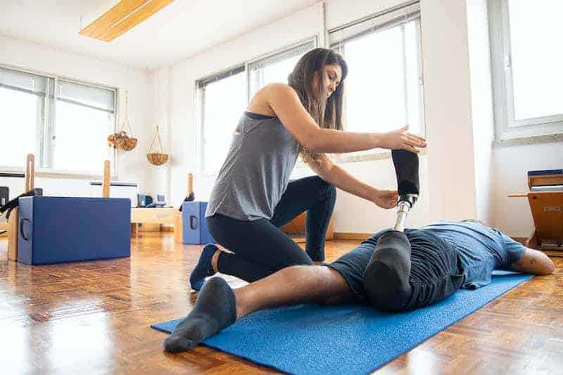 A woman is assisting a person who is lying face down on a blue exercise mat. She is holding and adjusting the person's prosthetic leg. The setting appears to be a physical therapy or rehabilitation room, with exercise equipment visible in the background.