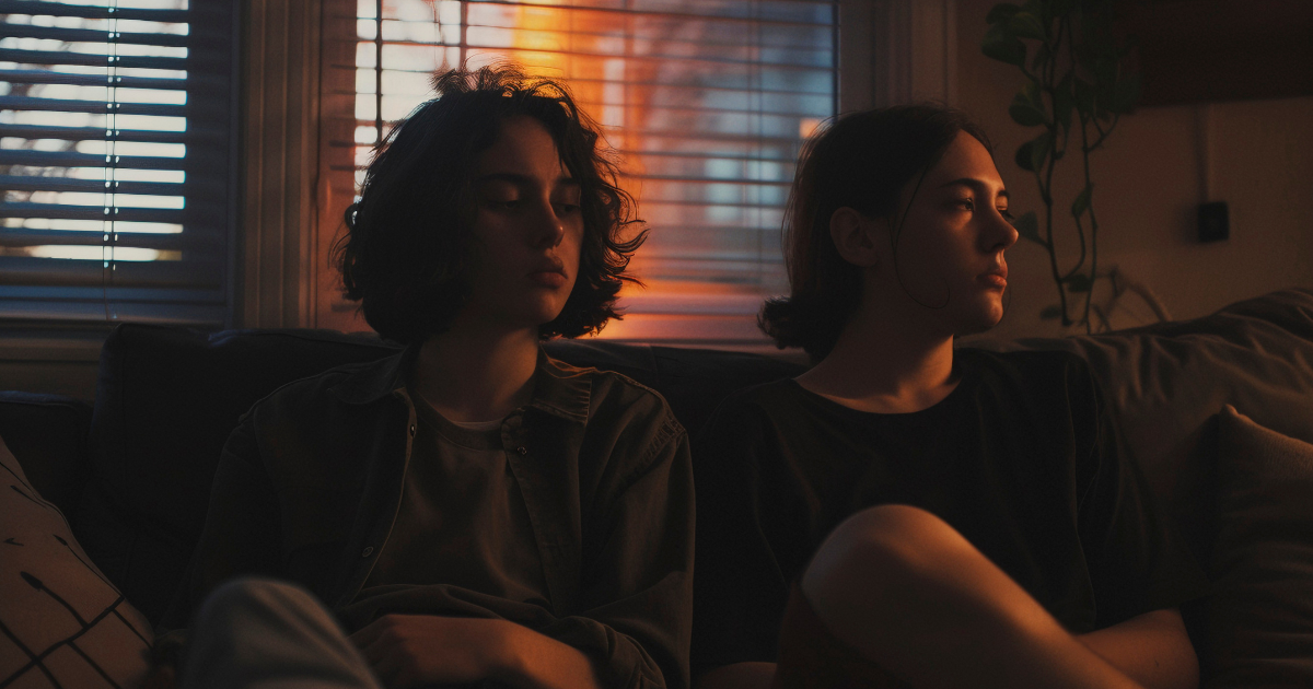 Two people sit in moody silence on a couch, avoiding eye contact as evening light filters through the blinds, capturing the quiet tension that can exist when values or temperaments clash without open understanding.