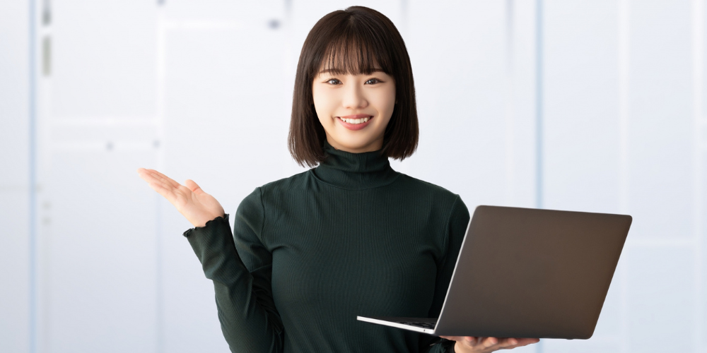 ノートパソコンを持ち、笑顔で手のひらを広げて案内するポーズの若い女性