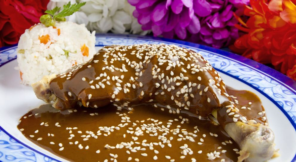 A imagem apresenta um prato de Mole Poblano, uma iguaria tradicional mexicana. Uma coxa de frango é coberta com um molho espesso e escuro, feito com especiarias e chocolate, e finalizada com sementes de gergelim. Ao lado, há arroz branco com legumes. Flores coloridas decoram o fundo, reforçando a cultura mexicana.