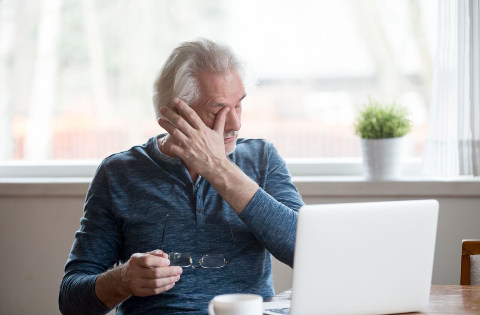 A man works on a computer and removes his glasses to rub his eyes due to dry eye symptoms.