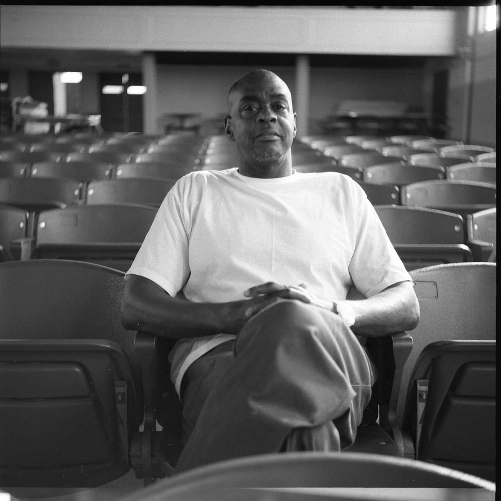 Image: Photograph of Mike Sullivan by Justin Jones, 2023. Mike, a middle-aged Black man with a closely-shaved head sits in a plastic chair in an auditorium with his legs crossed and his hands clasped in his lap. He looks at hte camera. Photo courtesy of Justin Jones.