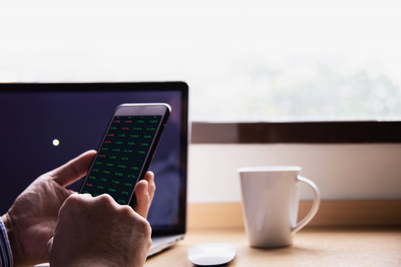 a person holding a smartphone displaying stock market data with green and red numbers, indicating financial performance or price changes with a cup and mouse on the table.