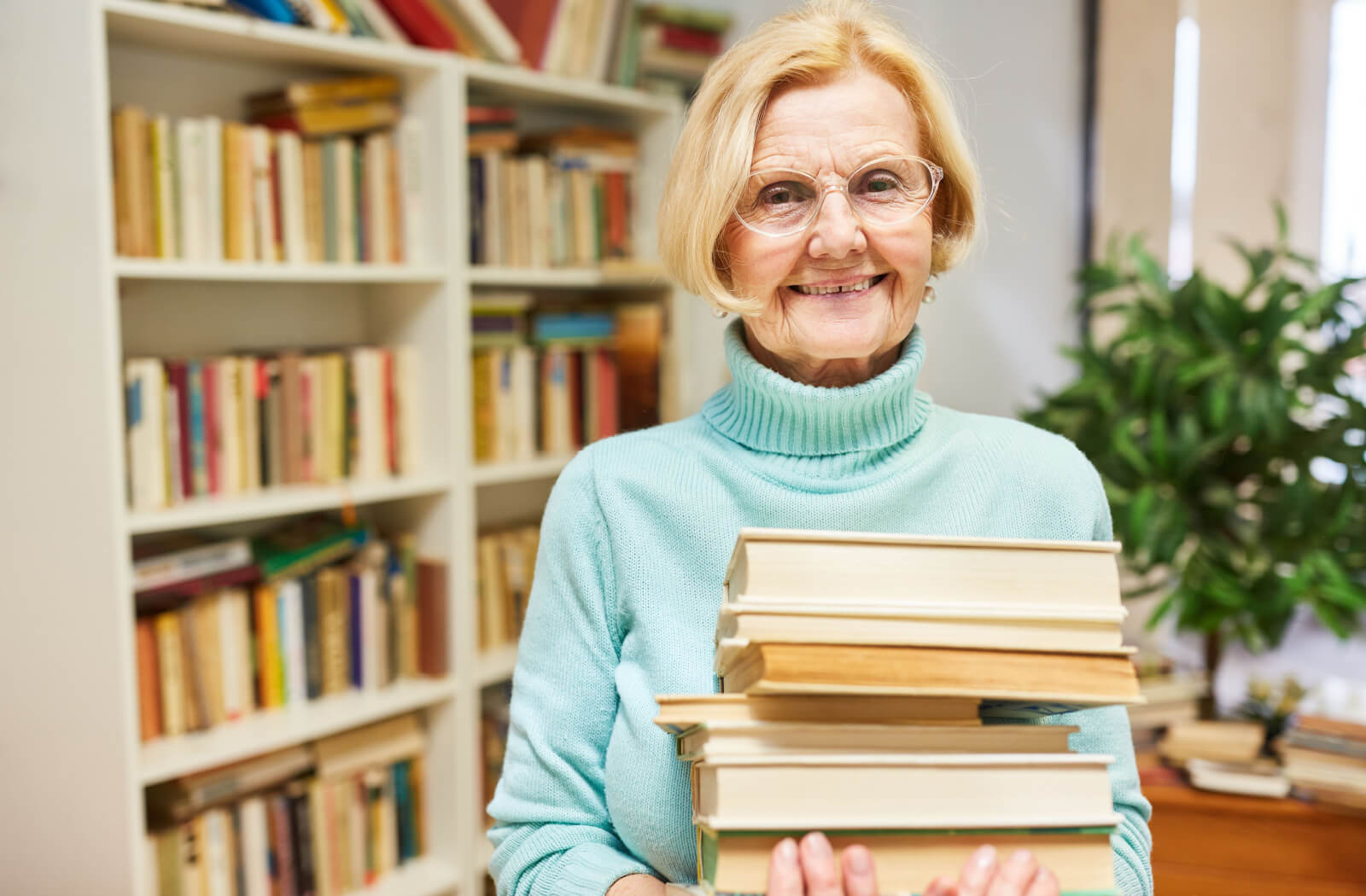 A smiling older adult in a library carrying a stack of books for their newest hobby in book club.