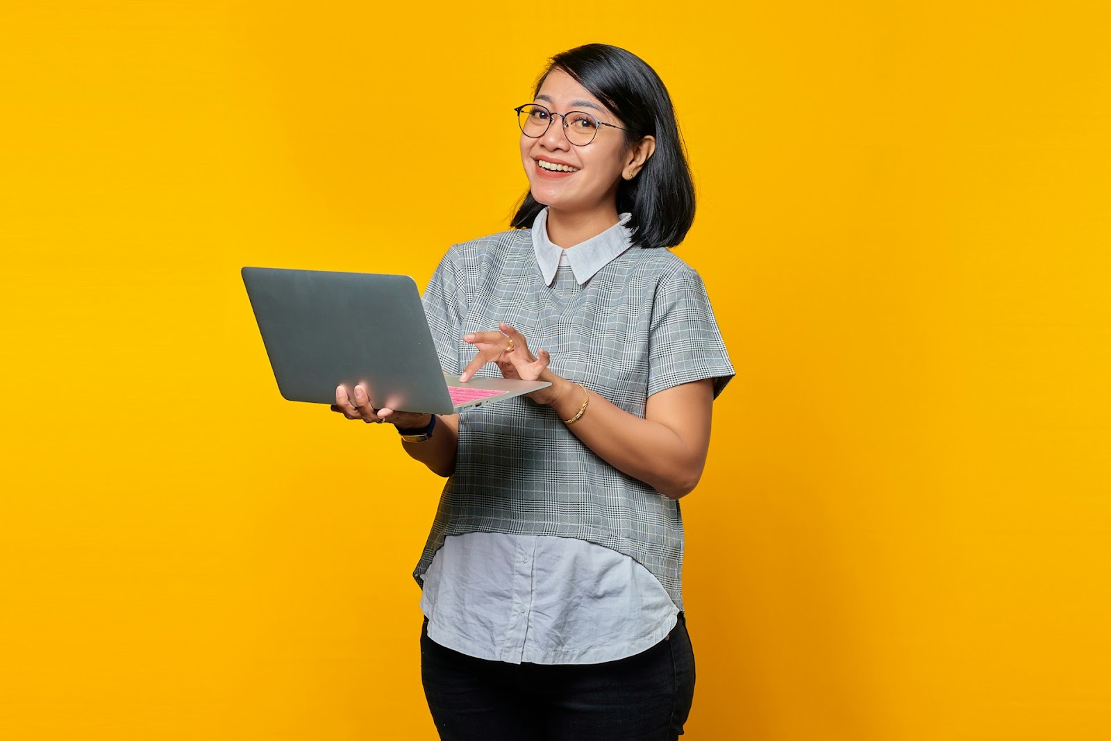 A woman wearing glasses smiles while holding a laptop against a bright yellow background.