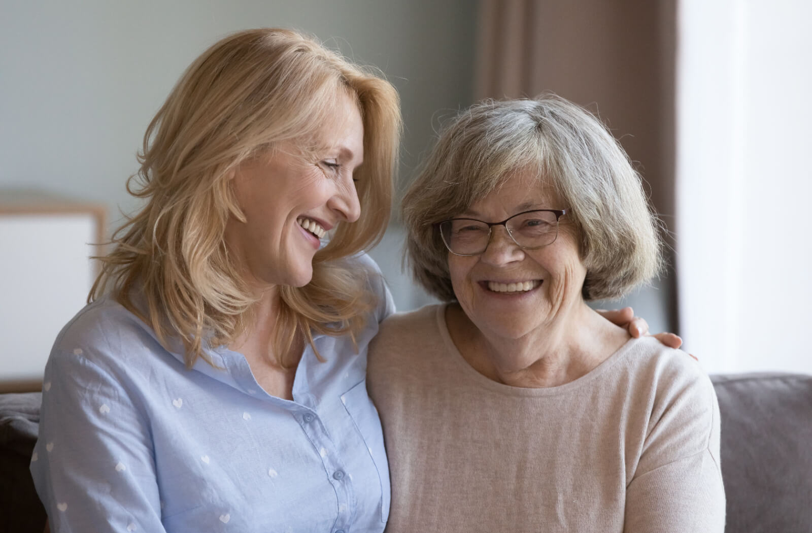 An adult visiting their older parent in a senior living community, smiling and hugging on the couch.