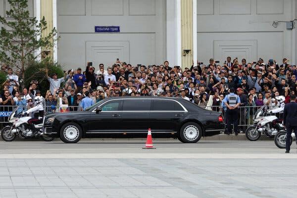 A black limousine flanked by white motorbikes drives past a crowd of people amassed behind a metal barrier.