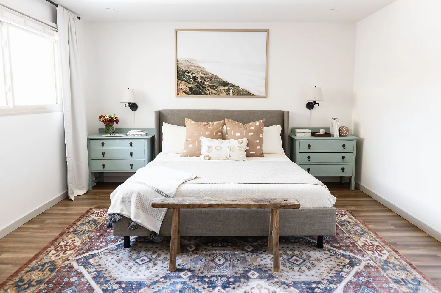 A modern, accessible bedroom with a gray upholstered bed frame, white bedding, and neutral-toned throw pillows. The room features mint green nightstands with black hardware, wall-mounted sconces, and a rustic wooden bench at the foot of the bed. A large framed coastal landscape print hangs above the bed, and a patterned area rug adds warmth to the wood flooring. Natural light filters in through a curtained window on the left.