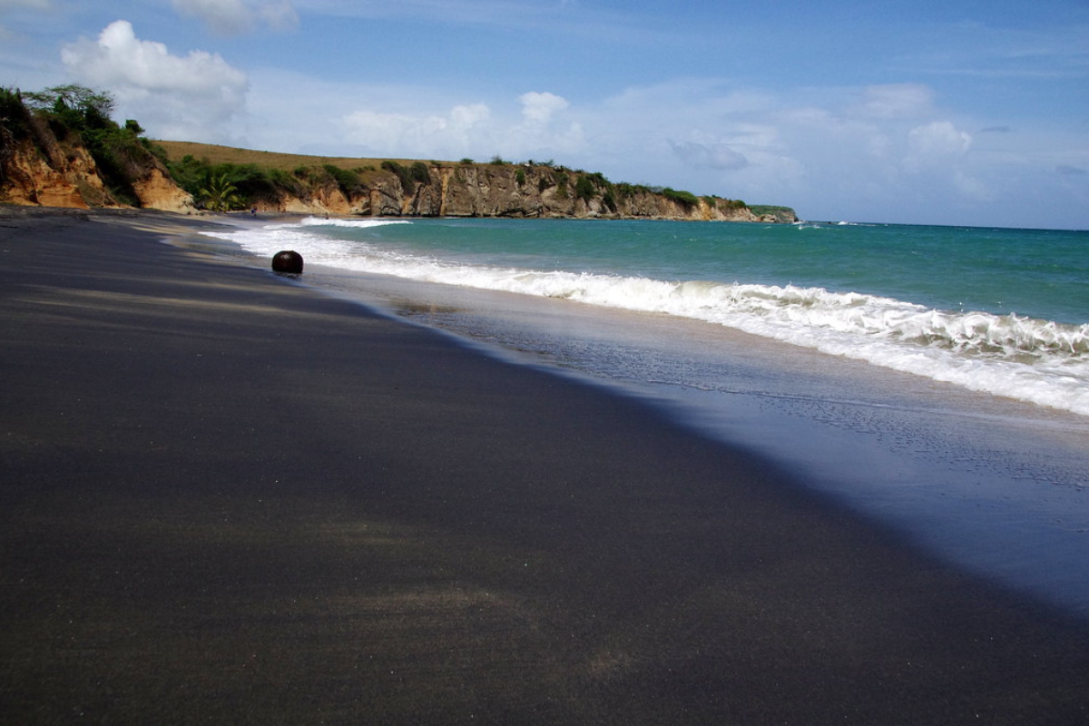 This is black sand beach and there is clean and clear water on the beach and there are big rocks near water