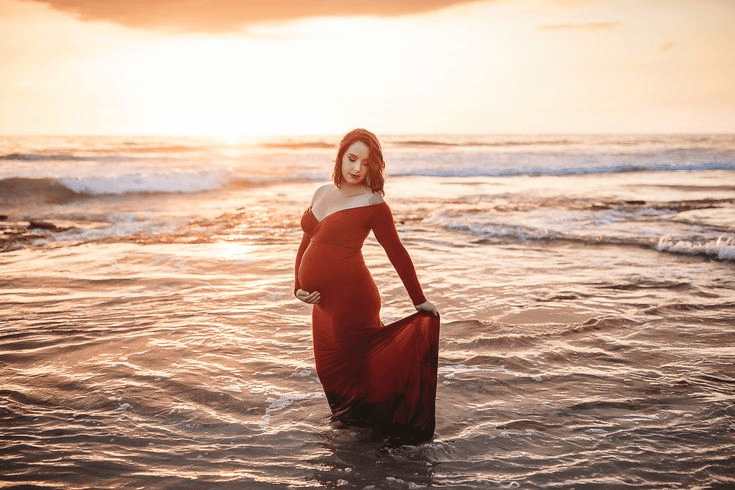 Maternity shoot with pregnant woman in red mermaid dress, captured standing in the ocean