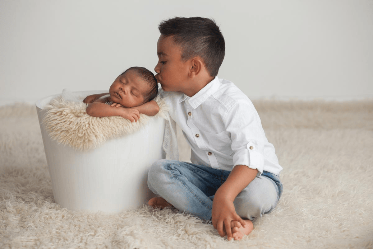 Baby photoshoot elder sibling softly kissing baby’s forehead capturing heartwarming moment.