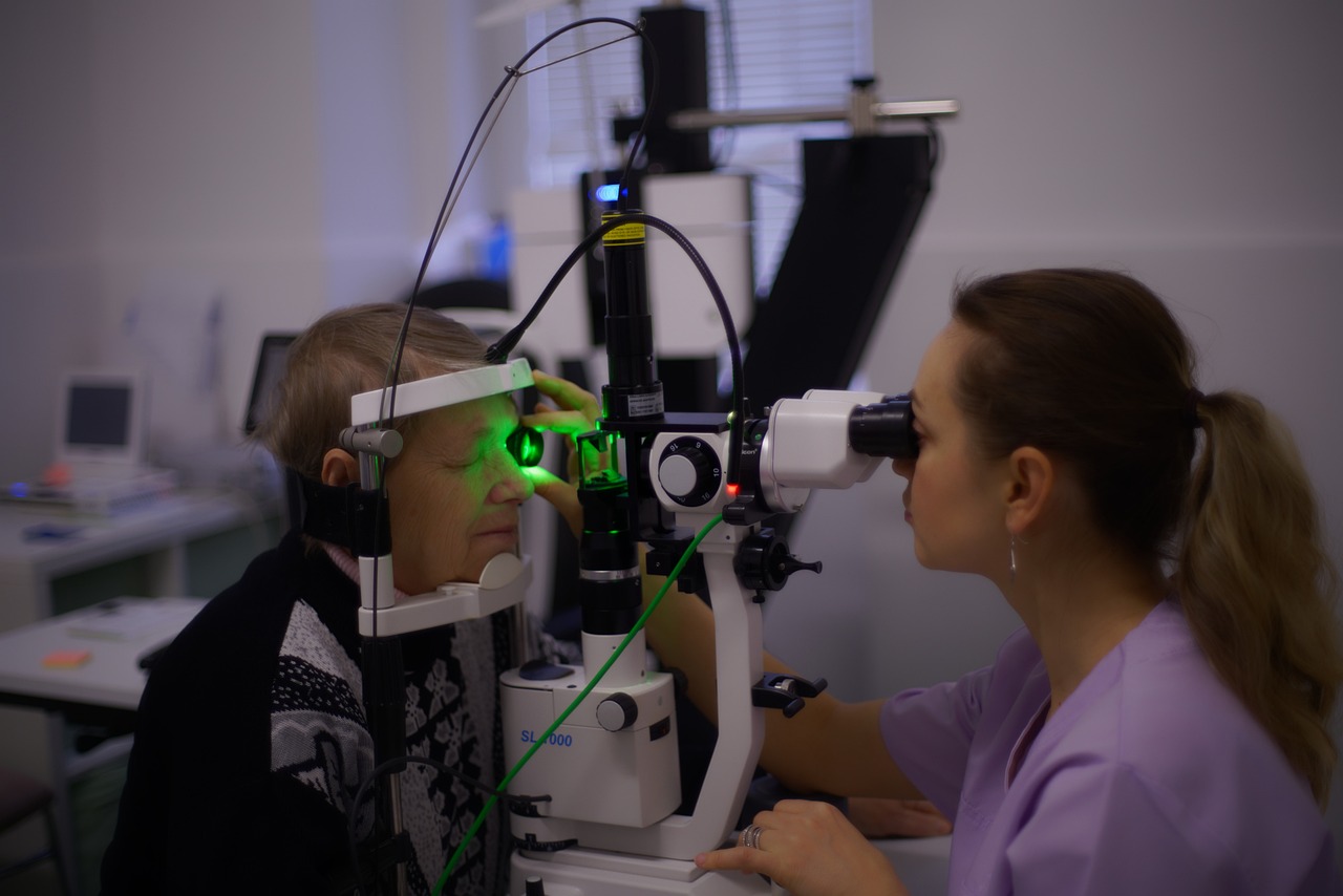 This image shows an optometrist  performing an eye examination on an elderly patient using a slit lamp. The optometrist, with her hair in a ponytail and wearing a light purple uniform, is looking through the device's eyepiece.