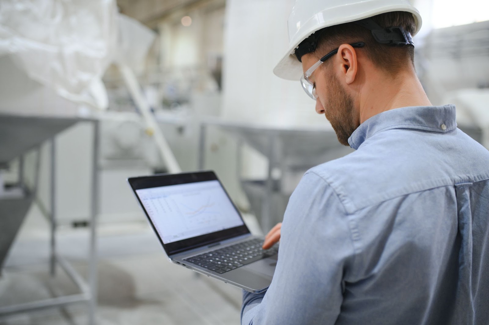 Side view of a technician or engineer wearing a headset, inputting data on a laptop while standing in an industrial factory, monitoring machinery and systems.